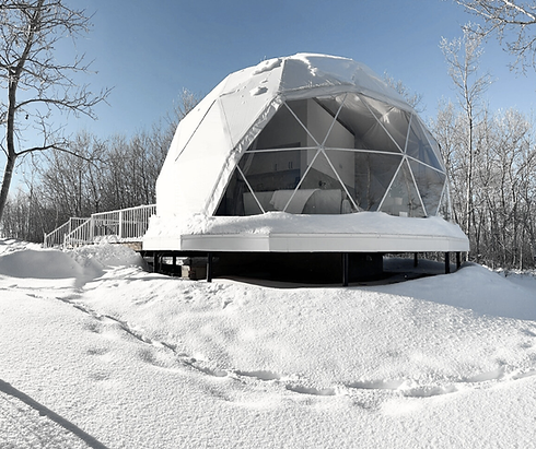 White geodesic dome covered in snow, surrounded by bare trees at Wanderlust Domes in Manitoba.