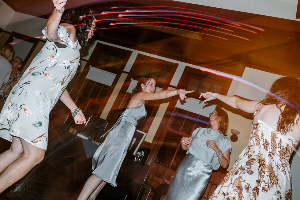 Women in floral dresses dance joyfully under colorful lights in a warmly lit room with wooden panels, creating a lively and festive atmosphere.
