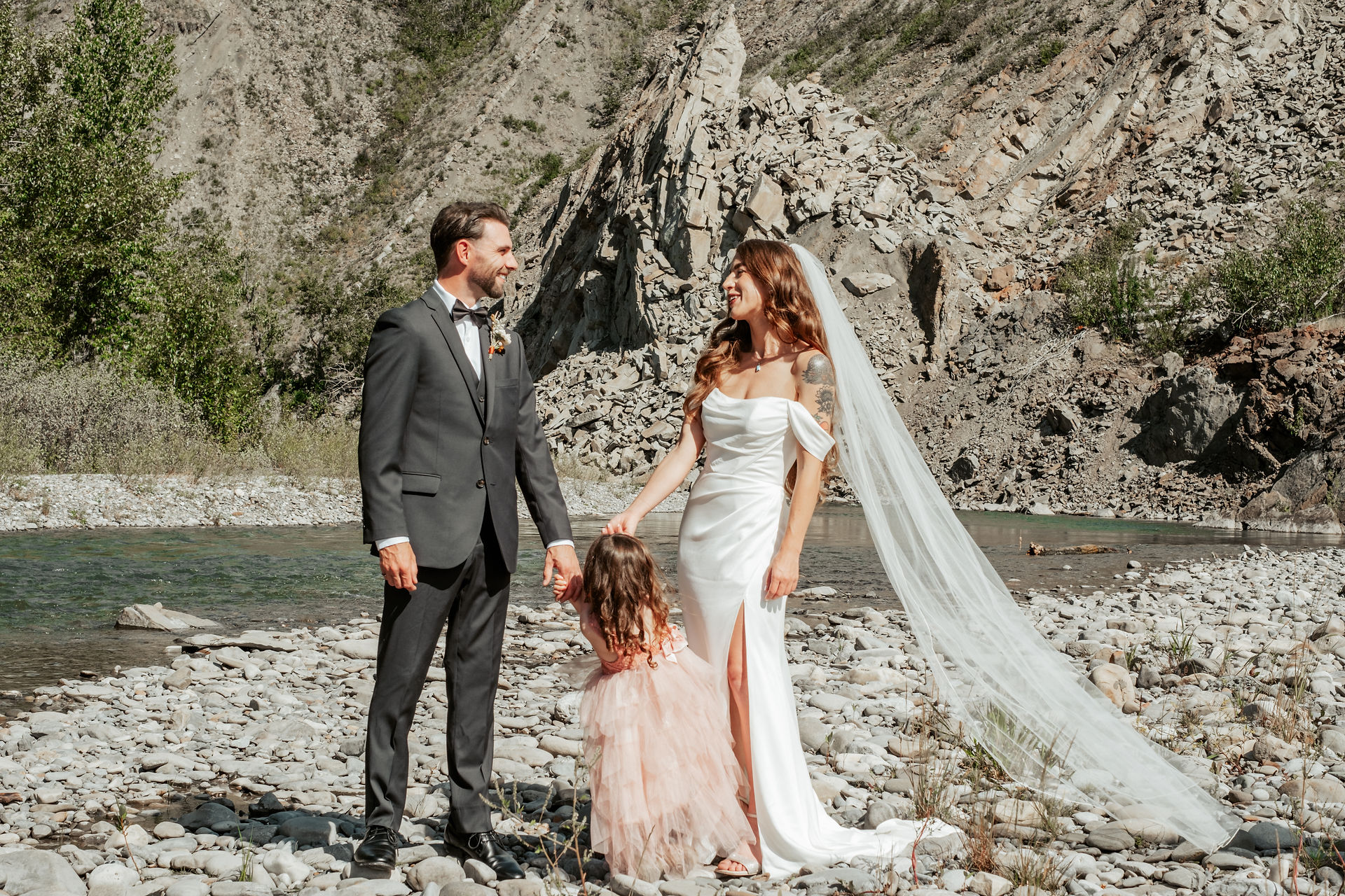 Bride and groom stand on a river bank with their daughter during the first look.