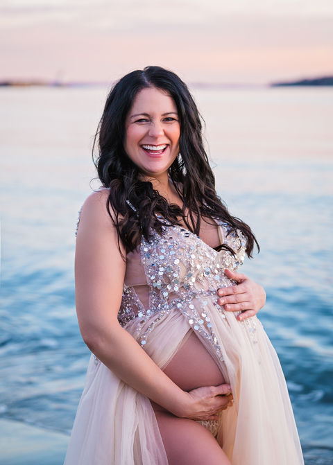 Beach Maternity Photography in Portland, Maine with photo of a pregnant woman on the rocky coast and beach at sunrise with bright blue water and soft, sparkly dress