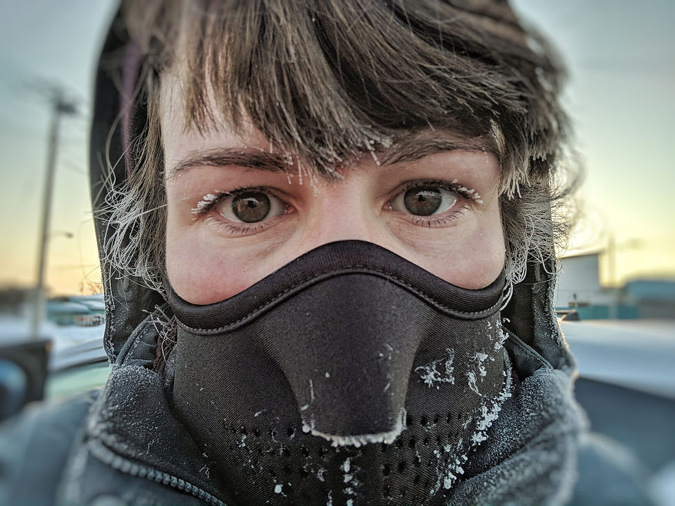 Person with frosty eyelashes and hair wears a black mask in a snowy outdoor setting. Background shows a pastel sky and power lines.