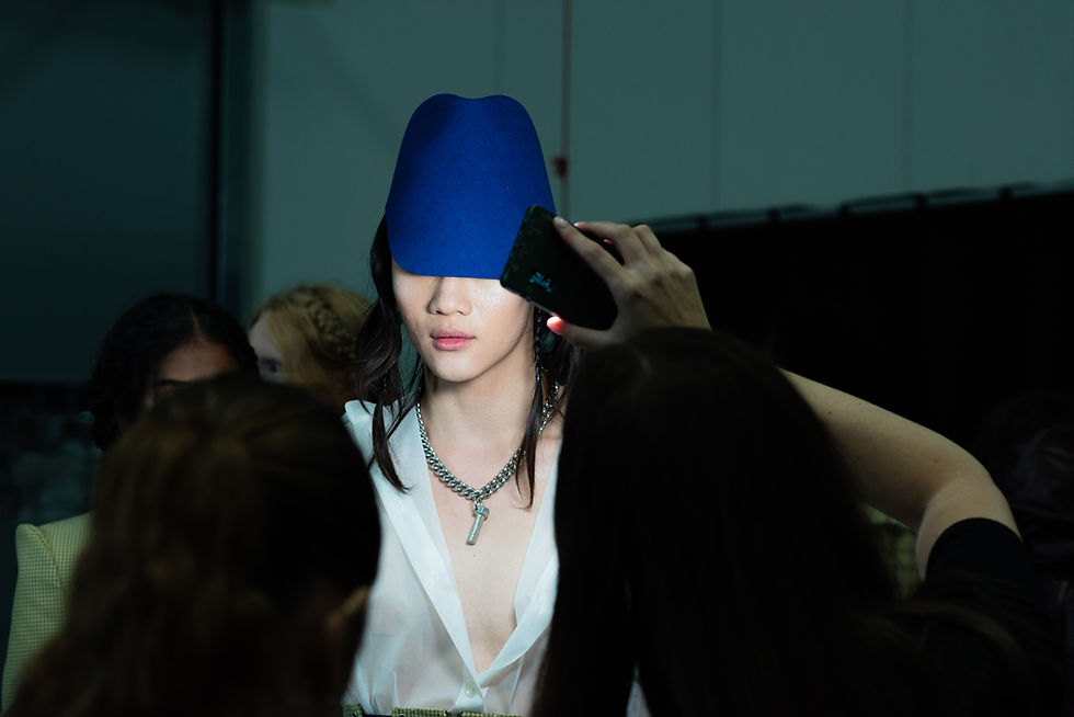 Fashion model in a blue hat and white blouse is surrounded by people and phones in a dim backstage room.