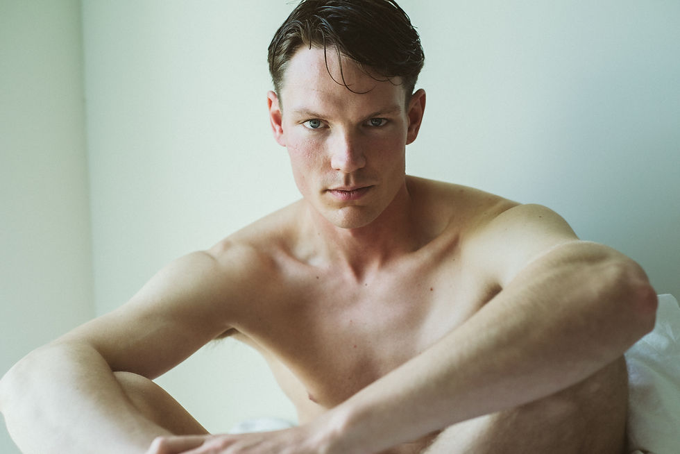 A shirtless person sits pensively against a light-colored wall, displaying a contemplative expression. Soft lighting enhances the calm mood.