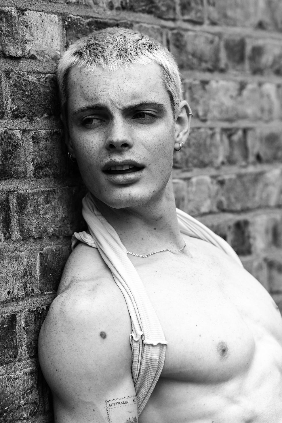 Black-and-white portrait of a shirtless person with short blond hair and earrings, leaning on a brick wall, pensive; AUSTRALIA tattoo.
