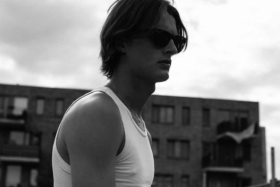 Black-and-white side profile of a young man in sunglasses and a white tank top, standing outdoors with blurred buildings.