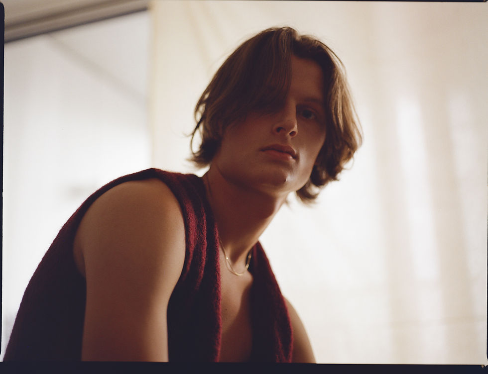 Portrait of a serious young person with shaggy hair in a burgundy top, lit softly against a bright plain background.