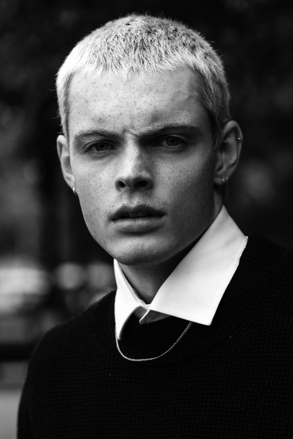 Black-and-white close-up portrait of a young man with bleached hair, earrings, and a serious expression against a blurred background.