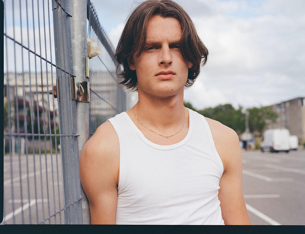 Young man in a white tank top leans by a metal fence outdoors, gazing seriously under a cloudy sky.