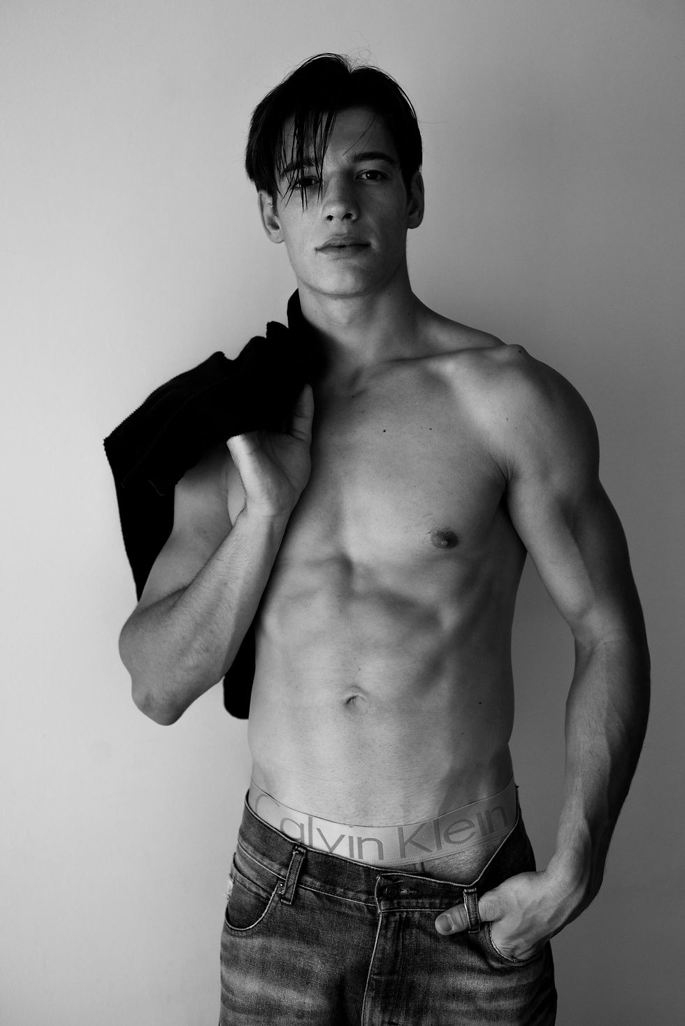Retrato en blanco y negro de un hombre torso desnudo con jeans Calvin Klein, sosteniendo una chaqueta negra, gesto serio.