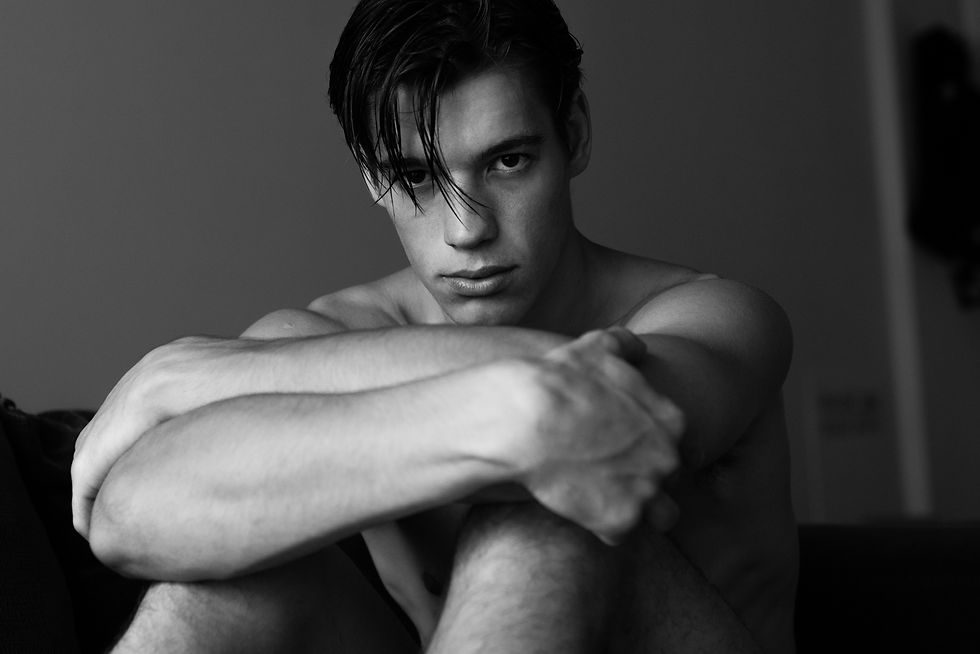 Retrato en blanco y negro de un joven sin camisa, abrazando sus rodillas, con mirada intensa y fondo gris.