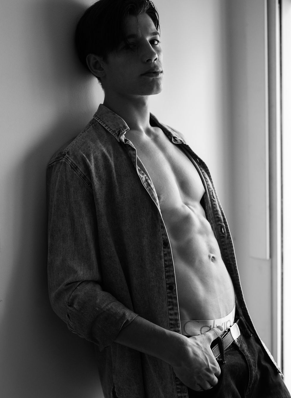 Joven posa en blanco y negro junto a una pared, con camisa vaquera abierta y torso desnudo; cintura Calvin Klein, gesto serio.