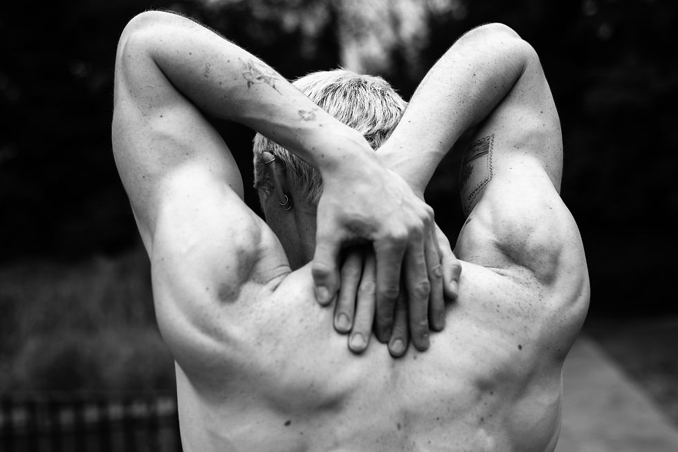 Black-and-white close-up of a shirtless person with short hair and tattoos, arms crossed behind their head outdoors.