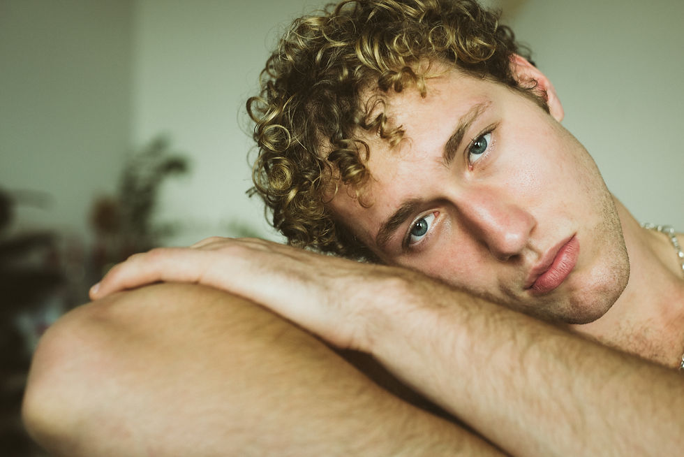 Close-up of a curly-haired young man resting on his arm, gazing at the camera in a softly lit indoor room.