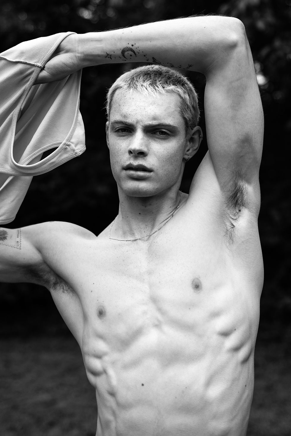 Black-and-white portrait of a shirtless young man with arm raised over his head, holding a shirt, outdoors, staring seriously.