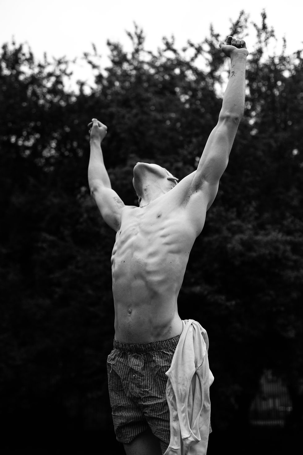 Black-and-white photo of a shirtless man outdoors, arms raised in triumph, with trees blurred in the background and a shirt draped at his side.