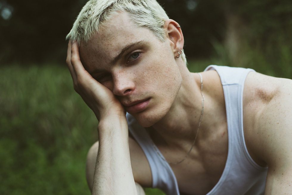 Close-up of a blond person in a white tank top, resting their face on a hand outdoors with a blurred green background, looking pensive
