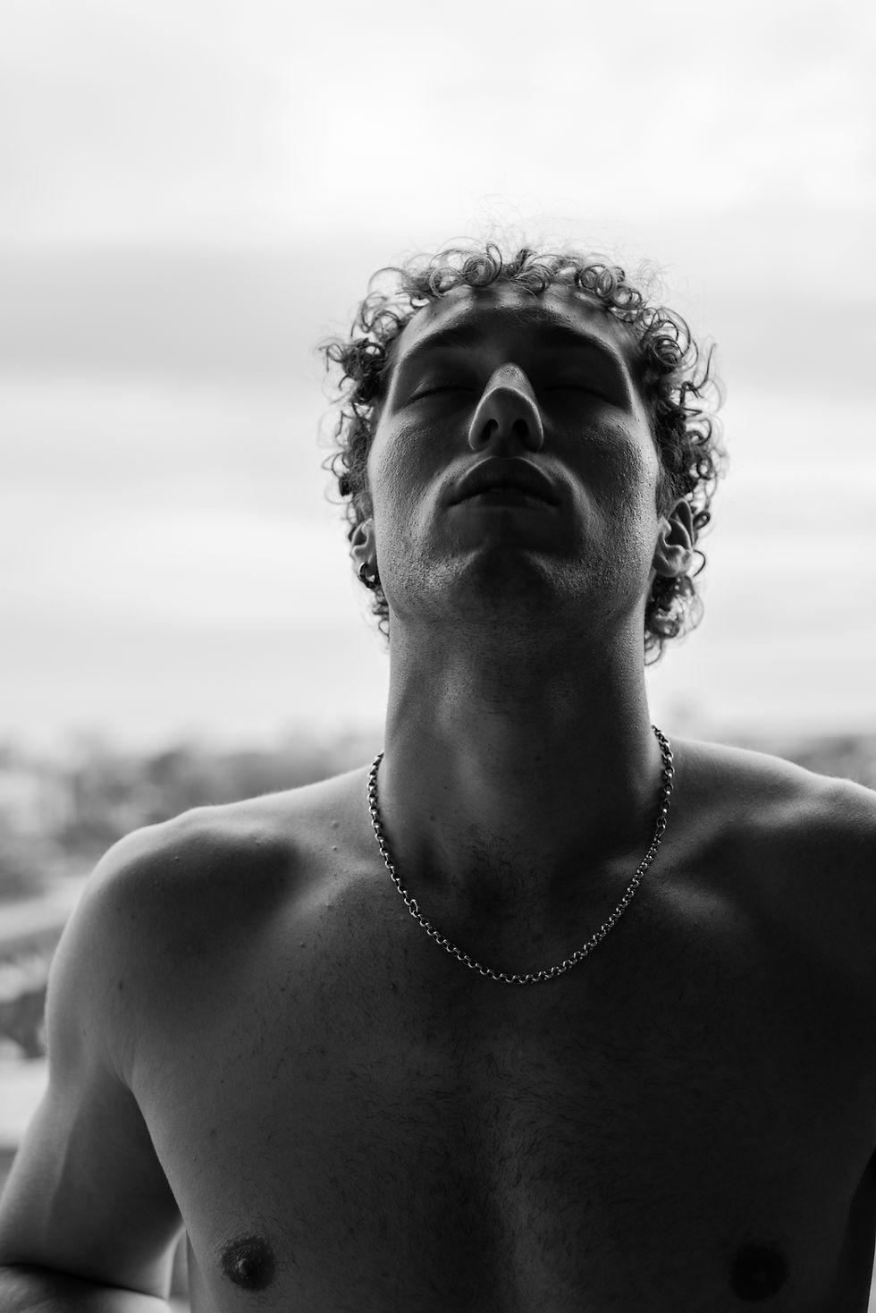 Black-and-white close-up of a shirtless man with curly hair and a chain necklace, eyes closed, against a bright outdoor sky.