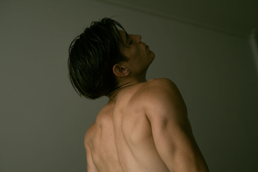 Joven sin camisa, de perfil y de espaldas, mira hacia arriba en una habitación tenue con luz verdosa.