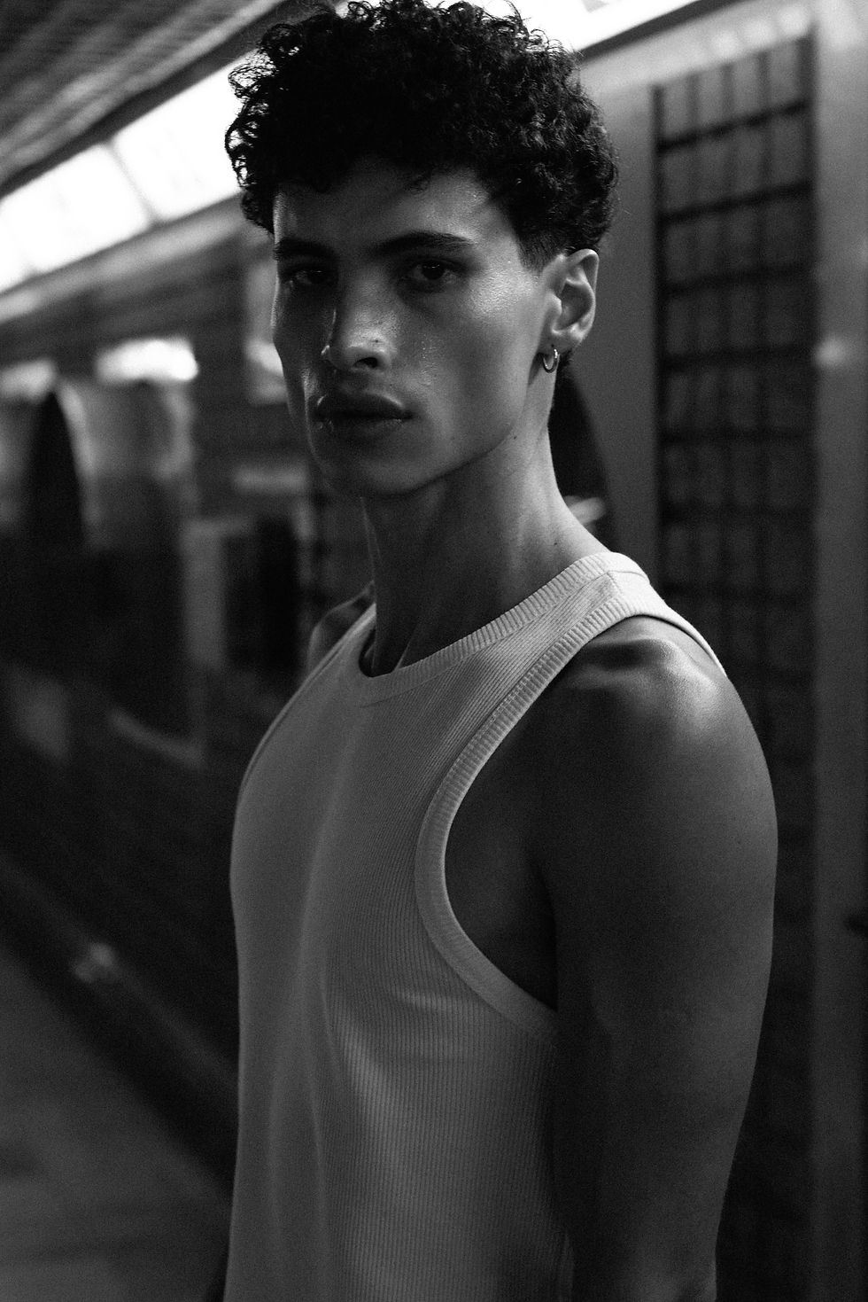 Close-up of Argentinian model Alejo in a white tank top, arms crossed, leaning against a red wall in the Milan subway