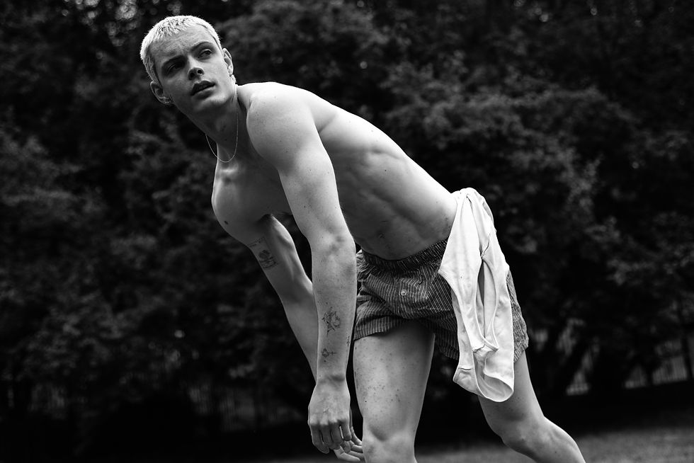 Black-and-white photo of a shirtless young man bending forward outdoors, wearing striped shorts with a white shirt tied at his waist, focused expression