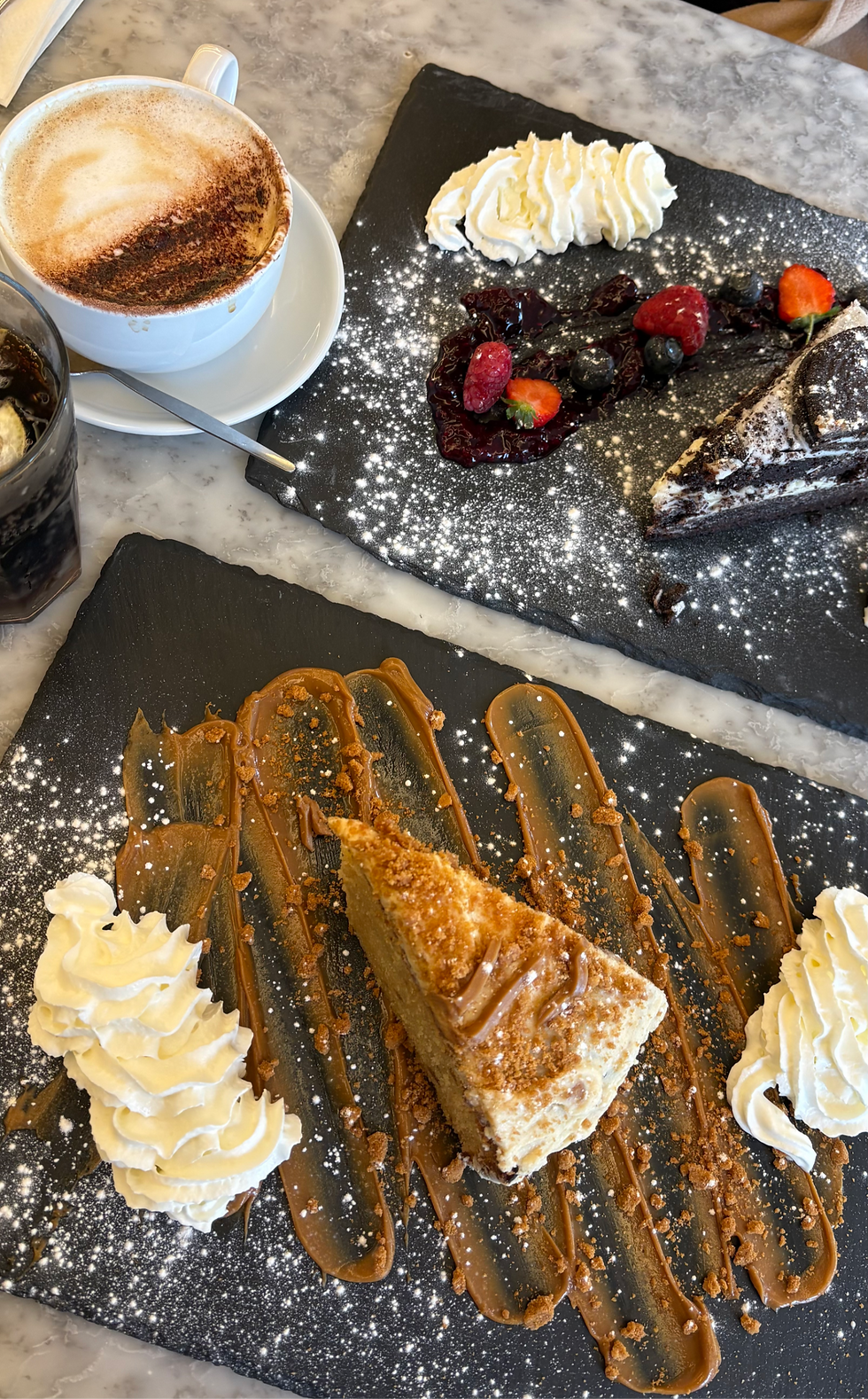 Coffee beside two plated desserts on black slate boards with whipped cream, berries, caramel, and powdered sugar on a marble table.