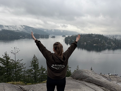 A person stands on a cliff with arms raised, overlooking a misty lake and forest. Cloudy sky. Jacket text reads "MANOAH ATHLETICS."