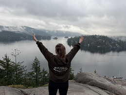 A person stands on a cliff with arms raised, overlooking a misty lake and forest. Cloudy sky. Jacket text reads "MANOAH ATHLETICS."