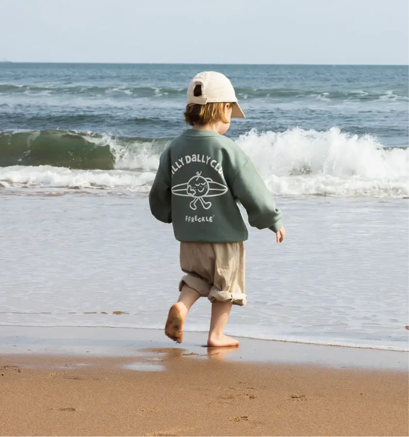 Child in a green sweater and beige hat walks barefoot on a sandy beach toward the ocean. Sweater reads "Dilly Dally Club Freckle." Waves in background.