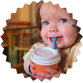 An image of a one year old child smiling at the camera drinking a babychino from an orange takeaway cup through a paper straw