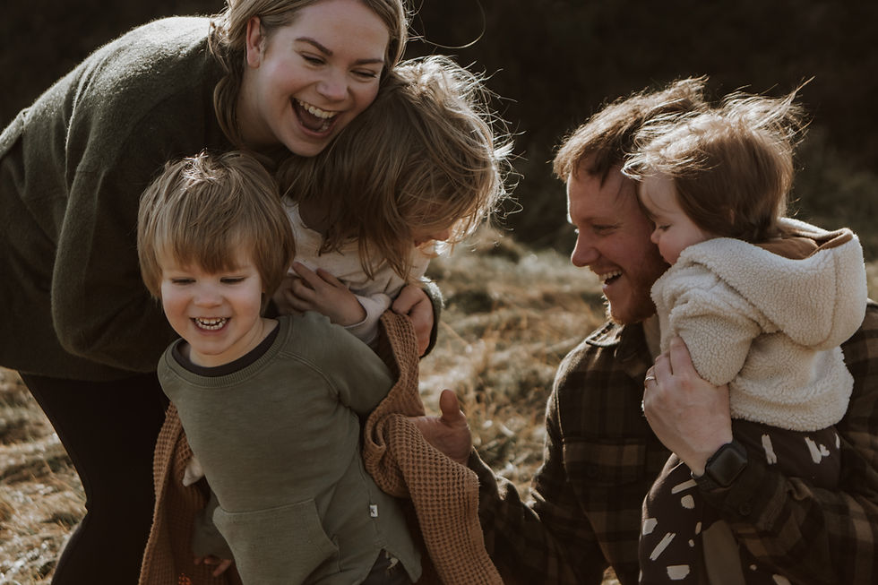 Family of four laughing and playing outdoors in a grassy field. The adults wear earthy tones, and the children are bundled in cozy clothing.