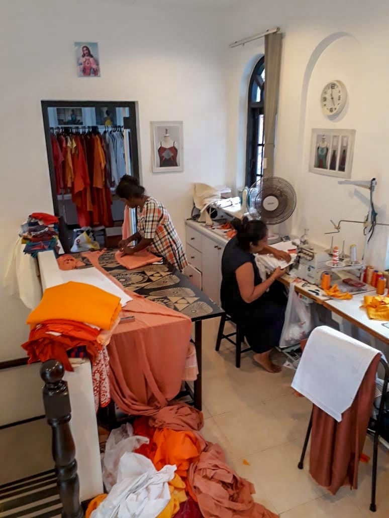 Two women making costumes. One woman on the right is sitting and working on a sewing machine. The woman on the left is standing and cutting patterns for sewing. There are costumes in the foreground.