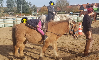 Ein Kind sitzt verkehrtherum auf einem braunen Pony und hat den Kopf auf dem Hintern des Ponys angelegt. Eine Frau führt das Pony dabei. Das Gesicht des Kindes wurde mit einem lächelndem Smiley zensiert.