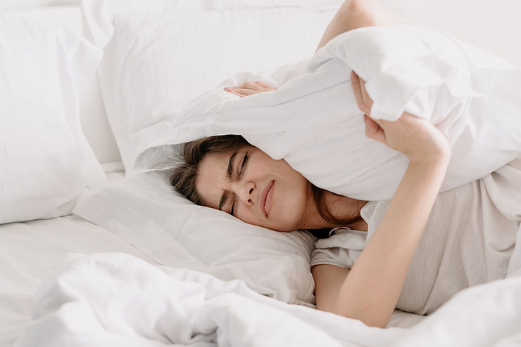 woman trying to sleep in bed with white sheets, covering her ears with a pillow to block out noise