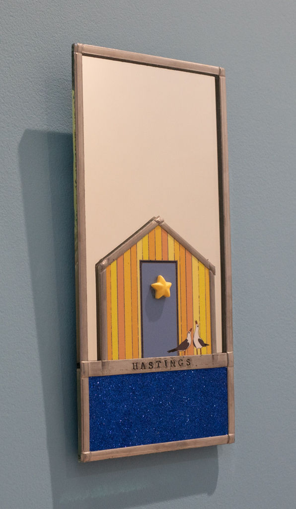 Indoor mirror with a yellow/orange panelled beach hut with text saying Hastings, hanging on wall background.