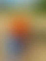 A woman in black and white plaid flannel wearing a large, orange backpack, green Tilley hat and sandals walking on a dirt road with houses in the background