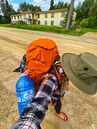 A woman in black and white plaid flannel wearing a large, orange backpack, green Tilley hat and sandals walking on a dirt road with houses in the background