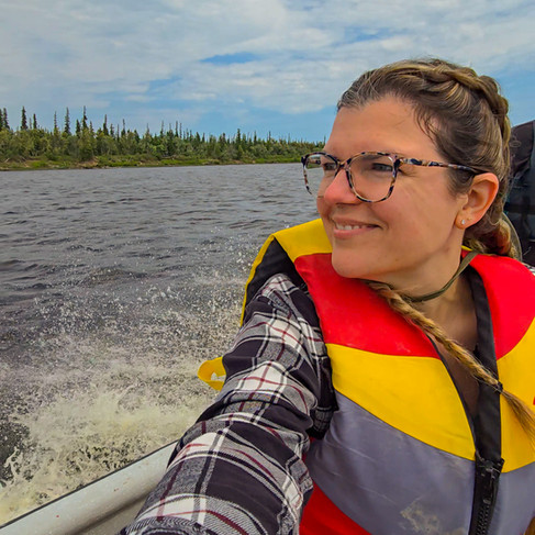 A smiling woman with dark brown braids sitting in a boat looking at the view. She is wearing a black and white plaid flannel shirt and a yellow, grey and red life jacket.