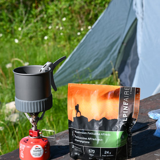 An MSR pocket rocket stove attached to a fuel canister with a pouch of Mushroom Fettucine, with a tent in the background