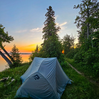 A gret sea to summit tent pitched on grass with trees and a river with the sun rising in the background