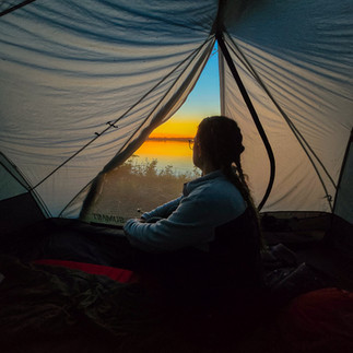 A woman with braided hair looking out of her grey tent with the sun setting over the river in the background