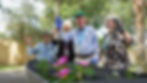A joyful outdoor scene showing four smiling residents waving to the camera while gardening on a sunny day. A gentleman in a green cap and light blue shirt stands in the center wearing blue gloves, flanked by two cheerful women and another seated gentleman, all gathering around a boat-shaped planter filled with bright pink and purple flowers with lush green trees in the background.