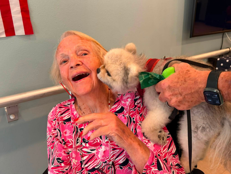 An elderly woman wearing a vibrant pink and black patterned top smiles brightly as a small, fluffy dog licks her cheek. A person's hands are visible holding the dog on a leash while wearing a black smartwatch on their wrist. In the background, a light green wall features an American flag hanging on the left and a silver handrail running horizontally behind the woman.