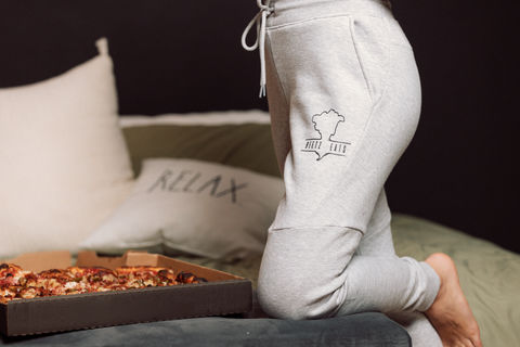 Model wearing gray sweatpants from a streetwear brand, lounging on a bed next to a pizza box.