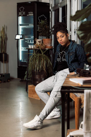 Person wearing gray joggers and a hoodie, seated casually in a modern café with plants in the background.