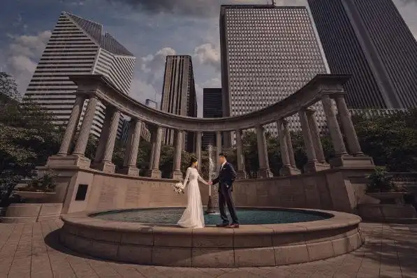 "Bride and groom hold hands at the center of a circular stone fountain, with Chicago's towering skyline stretching above, embodying the urban elegance captured by Studio Soo's wedding photography expertise."