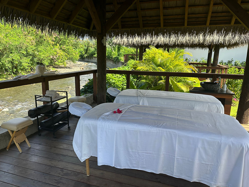 Two massage tables set up in a wooden cabana with a thatched roof, part of the Red Lane Spa at Sandals St. Vincent. A small river flows nearby, surrounded by tropical foliage.