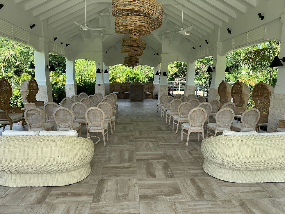 Interior view of the open-air Riverside Pavilion at Sandals St. Vincent, featuring white chairs arranged for a ceremony, woven wicker seating, and decorative light fixtures. Lush greenery is visible in the background
