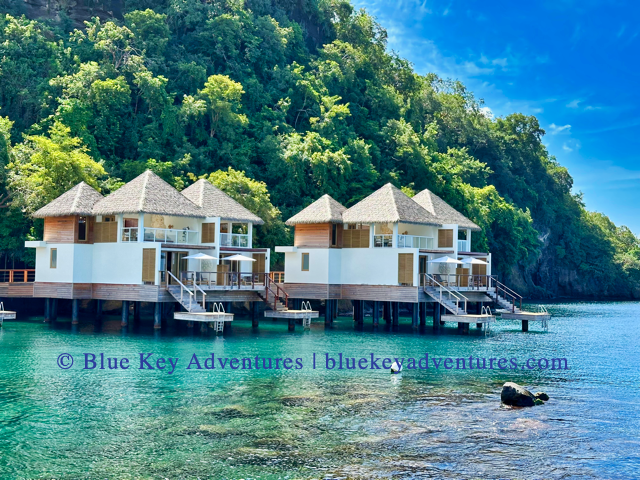 Two modern overwater two story villas with white and wooden exteriors, topped with peaked, thatched roofs, stand on stilts in clear, turquoise water. Each bungalow has a private wooden deck with steps leading down to the water. A lush, green hillside covered in trees rises steeply behind the bungalows, contrasting with the bright blue sky.