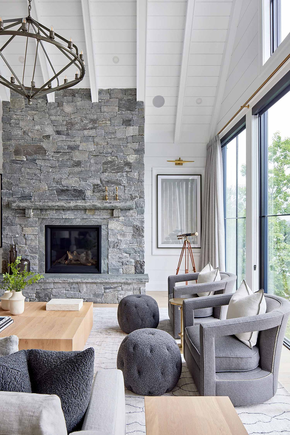 A bright and modern transitional style Muskoka great room interior. A dry stack stone fireplace with stone hearth and mantle grounds the room, with 12" white shiplap walls and white oak flooring. Expansive walls of black windows and earthy grey and taupe interior furnishings