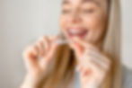 A smiling woman with long blonde hair holds a clear dental aligner between her fingers